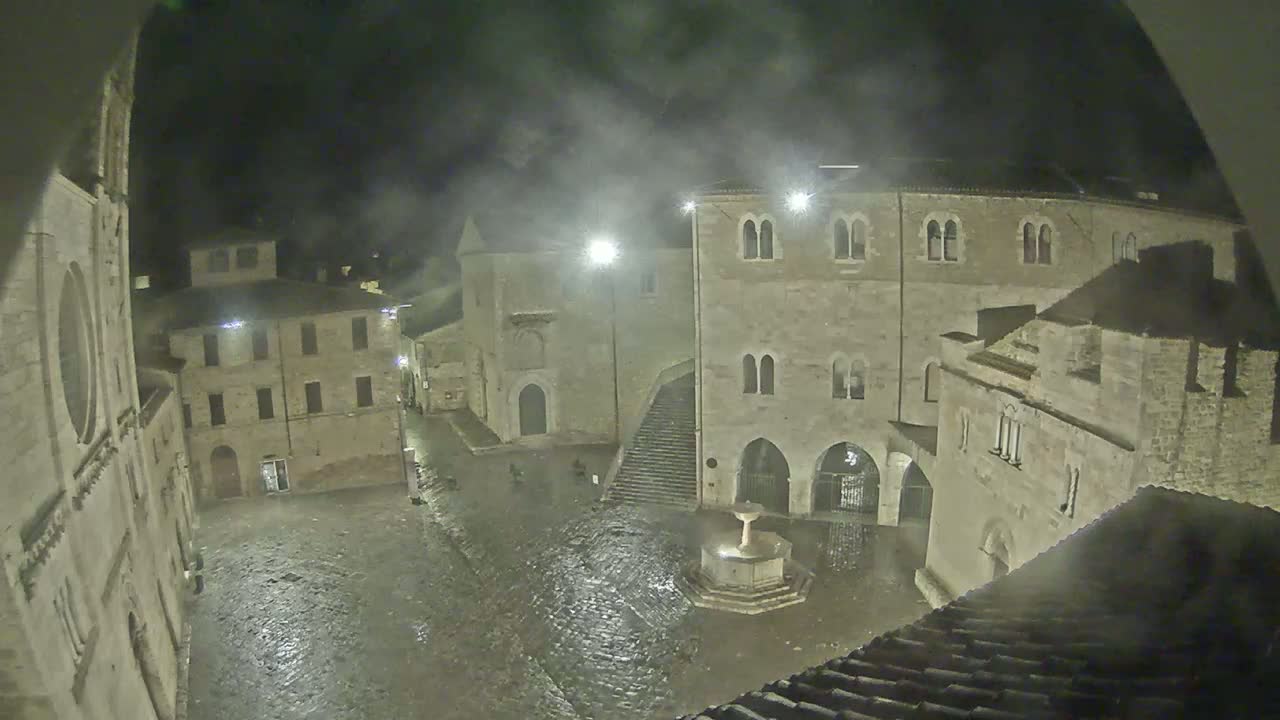 A nighttime view of a rain-soaked town square, surrounded by old stone buildings, with a fountain at its center.