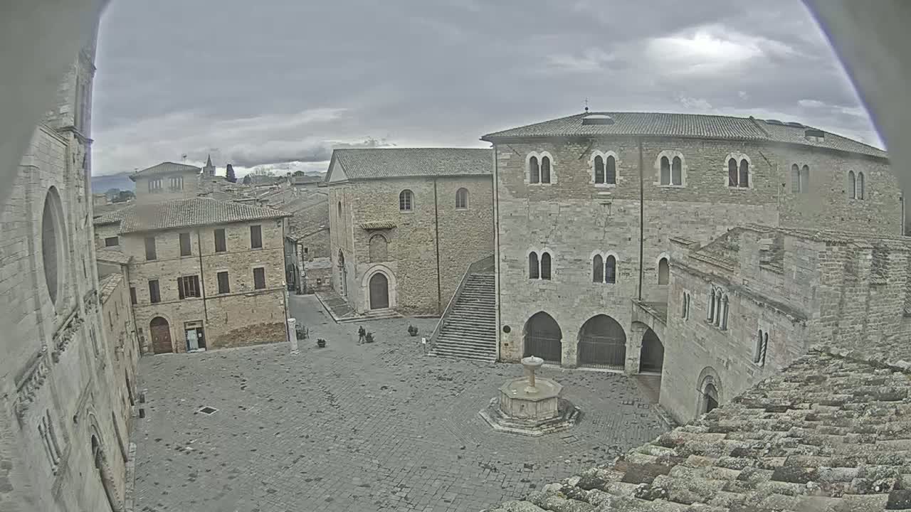 A stone plaza, under an overcast sky, is surrounded by medieval-style buildings, with a fountain in the center.