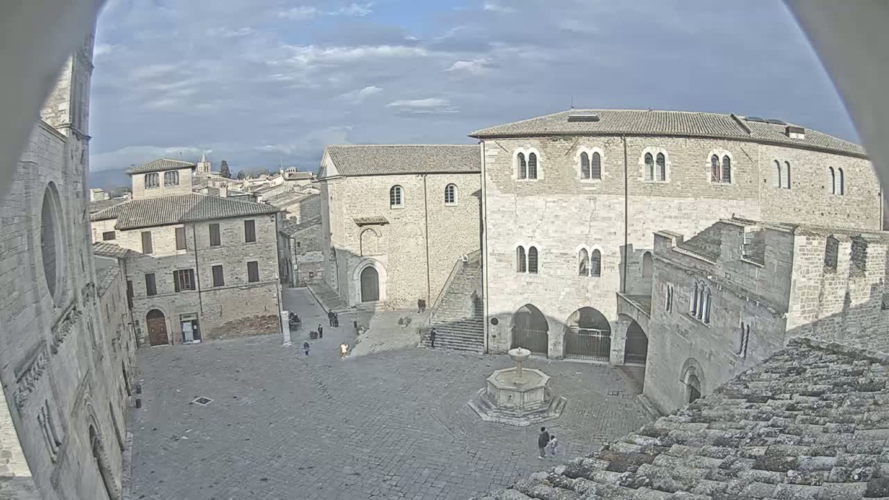 A mostly cloudy day reveals a cobblestone square surrounded by aged stone buildings, one featuring a central fountain, in what appears to be a European town.