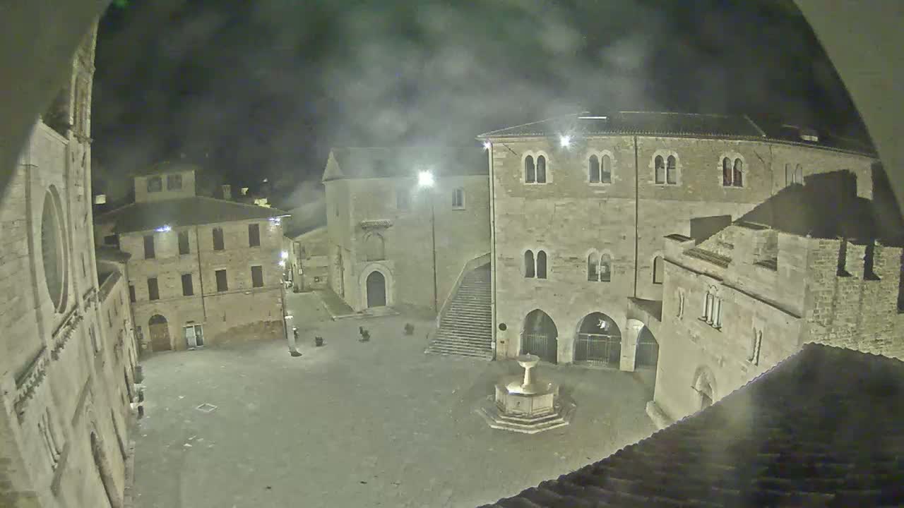 A nighttime view of a stone plaza with a fountain, surrounded by old buildings under a hazy sky.