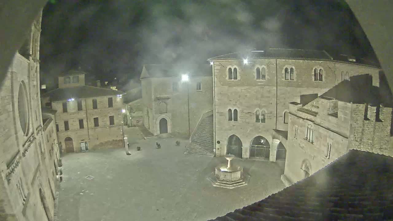 An empty town square at night, surrounded by old stone buildings under a hazy sky, features a central fountain.