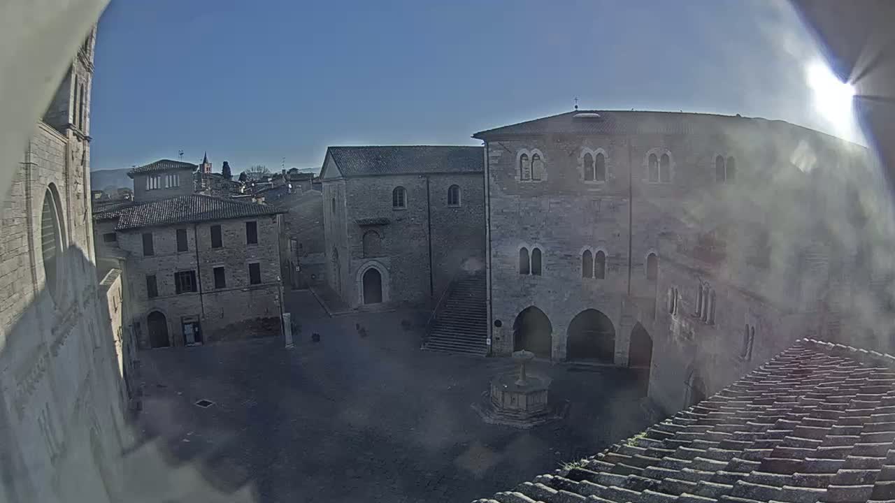 A sunny day reveals a high-angle view of a town square surrounded by aged stone buildings, featuring a central fountain.