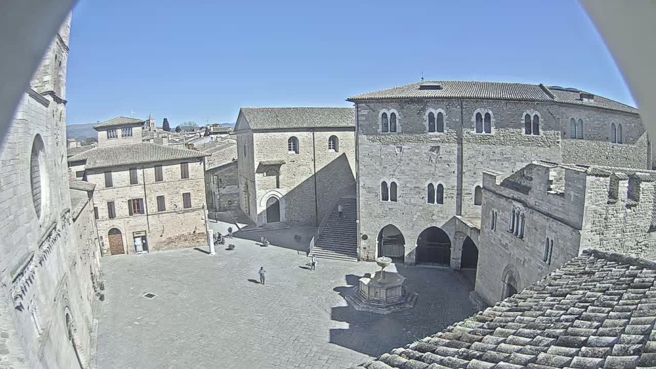 A high-angle view shows a stone plaza on a sunny day, surrounded by medieval-style buildings, with a fountain at the center.