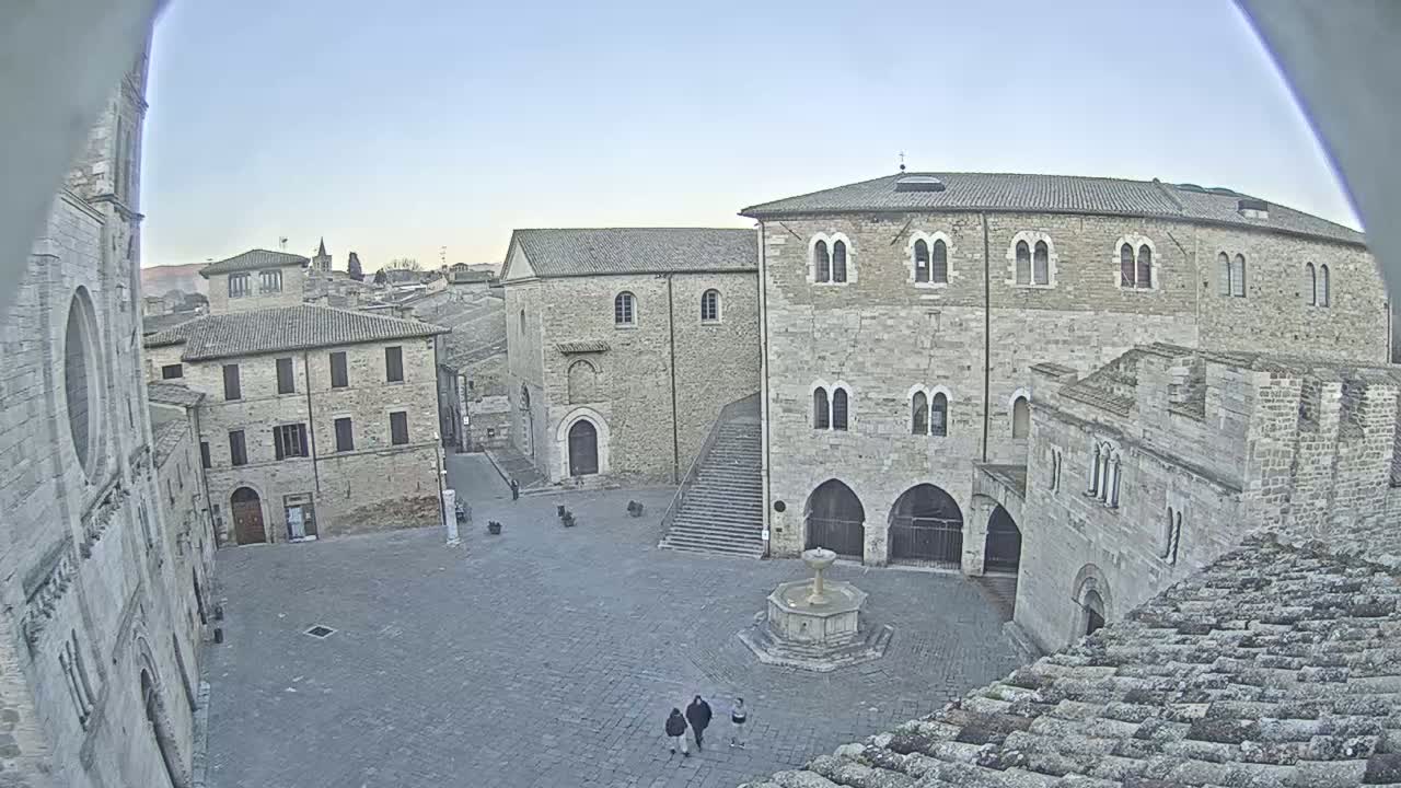 A stone plaza in a historic town is seen on a clear, bright day, featuring a fountain and several stone buildings surrounding it.