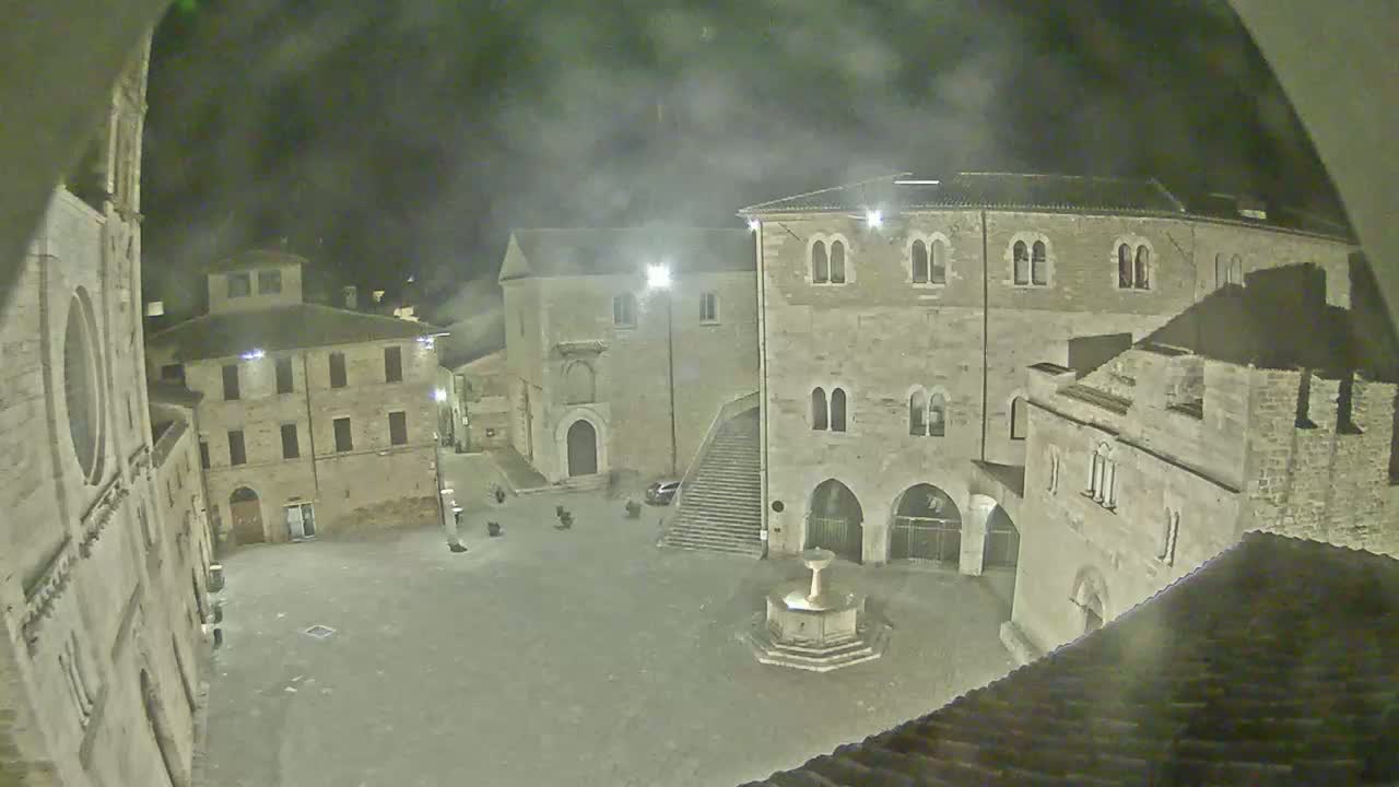 A nighttime view of a stone town square, featuring several aged buildings surrounding a central fountain, under hazy or foggy conditions.