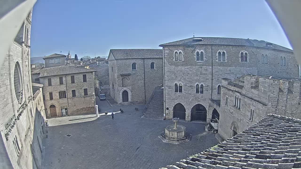 A mostly empty cobblestone town square is surrounded by several stone buildings under a clear blue sky.
