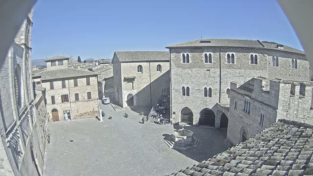 A high-angle view of a town square, under sunny skies, surrounded by medieval stone buildings, with a group of people gathered on some stairs.