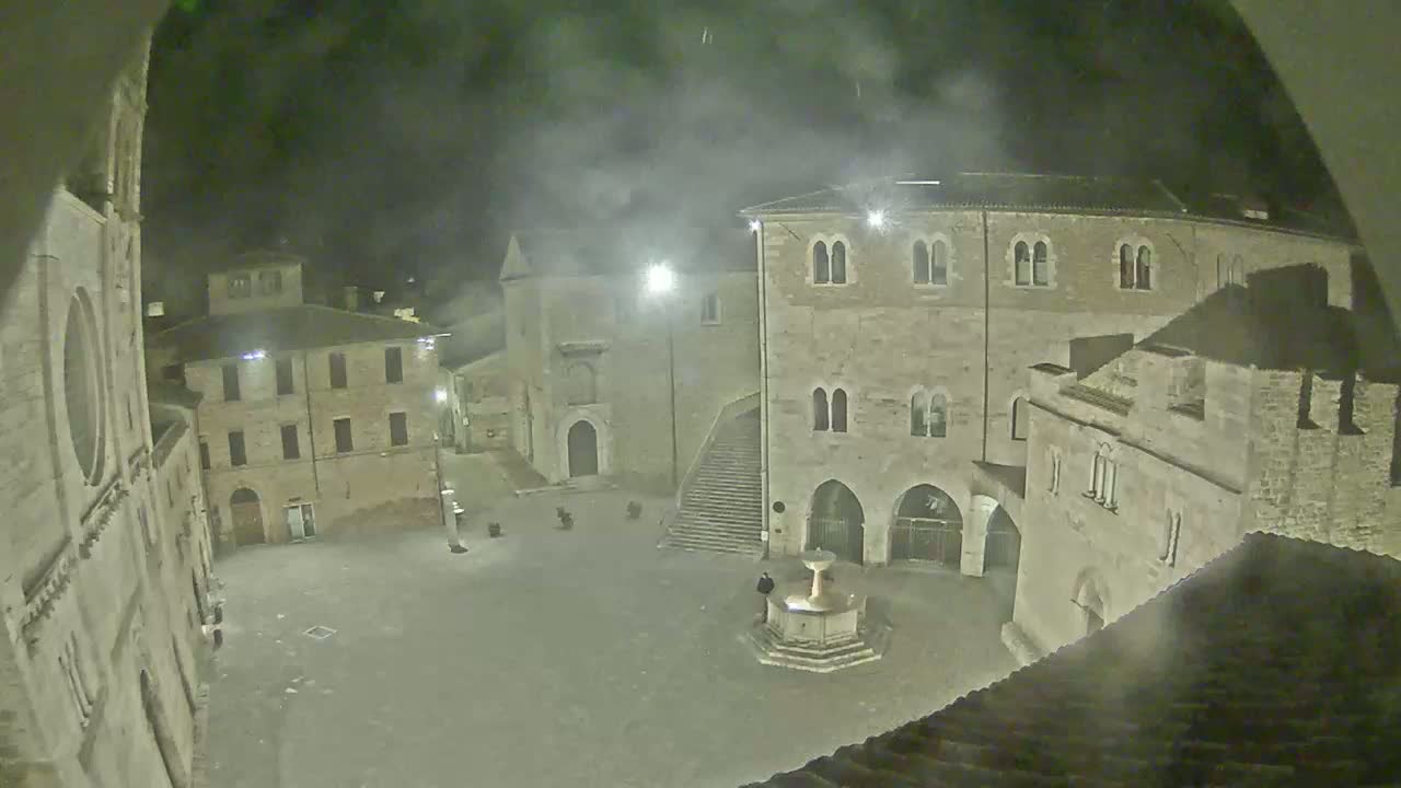 A nighttime view of a town square, surrounded by stone buildings, with a fountain in the center and a hazy or foggy atmosphere.