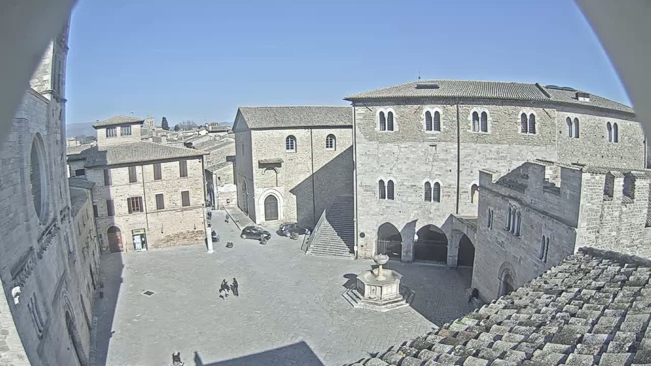A sunny day reveals a large, paved town square surrounded by old stone buildings, including a large stone structure with arched entryways, a fountain in the center, and a few people walking around.