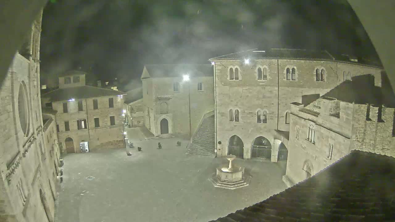 A nighttime, hazy view of a town square with stone buildings surrounding a fountain.