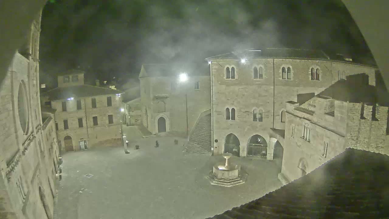 A nighttime view of a town square, partially obscured by fog or mist, features stone buildings surrounding a central fountain.