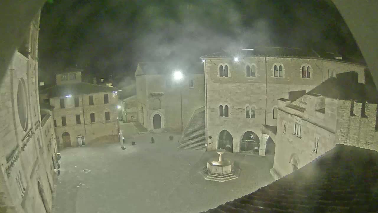 A nighttime, hazy view of a town square with stone buildings and a fountain in the center.