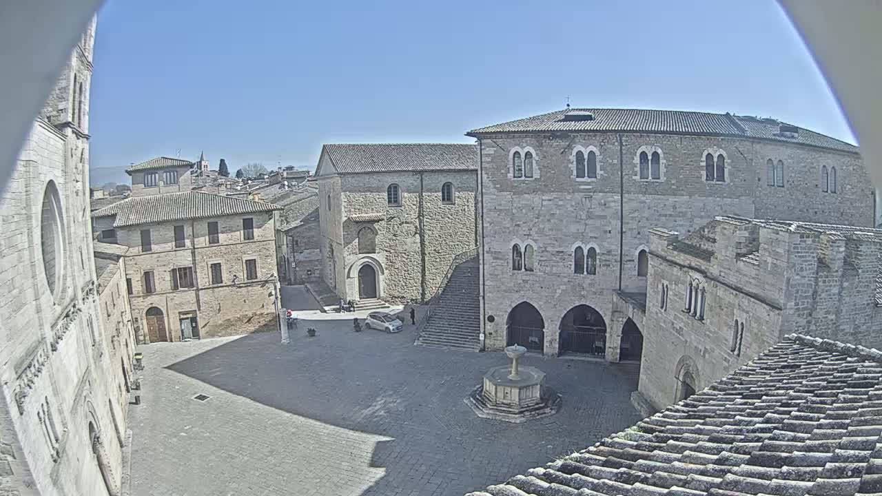 A high-angle view of a town square on a sunny day, surrounded by medieval-style stone buildings and a central fountain.