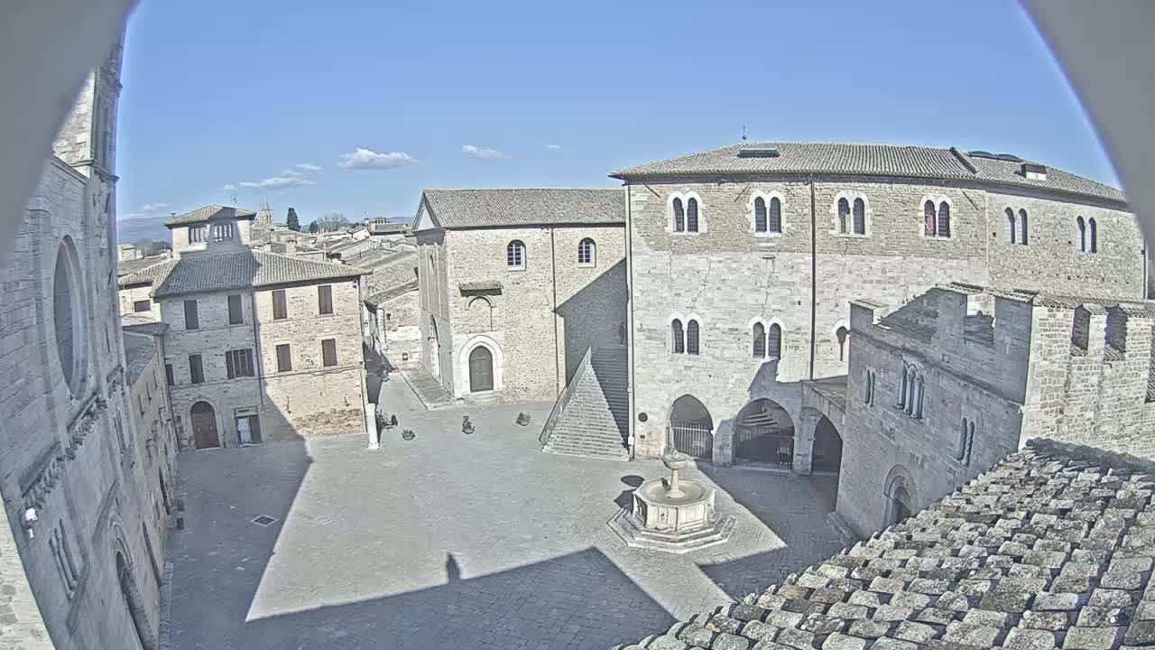 A high-angle view shows a town square in bright, sunny weather, surrounded by medieval-style buildings, with a fountain at its center.