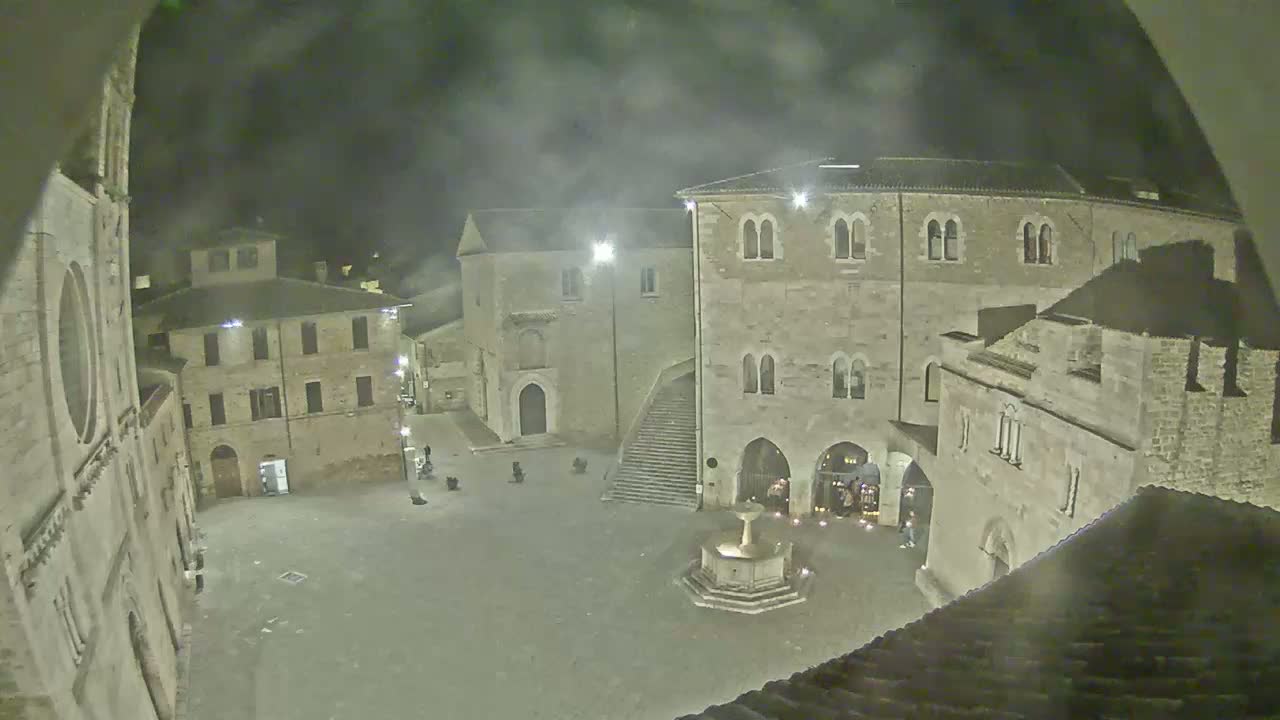 A nighttime view of a town square with stone buildings surrounding a fountain, under hazy or foggy conditions.