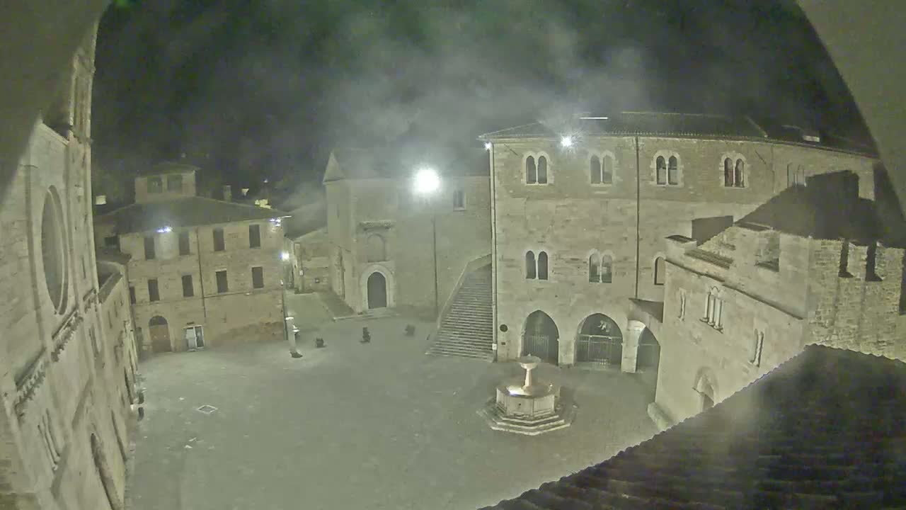 A nighttime view of a town square, featuring stone buildings and a fountain, under a hazy sky.