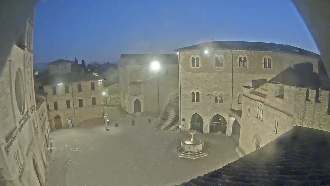A twilight view of a town square, centered on a fountain, surrounded by stone buildings.