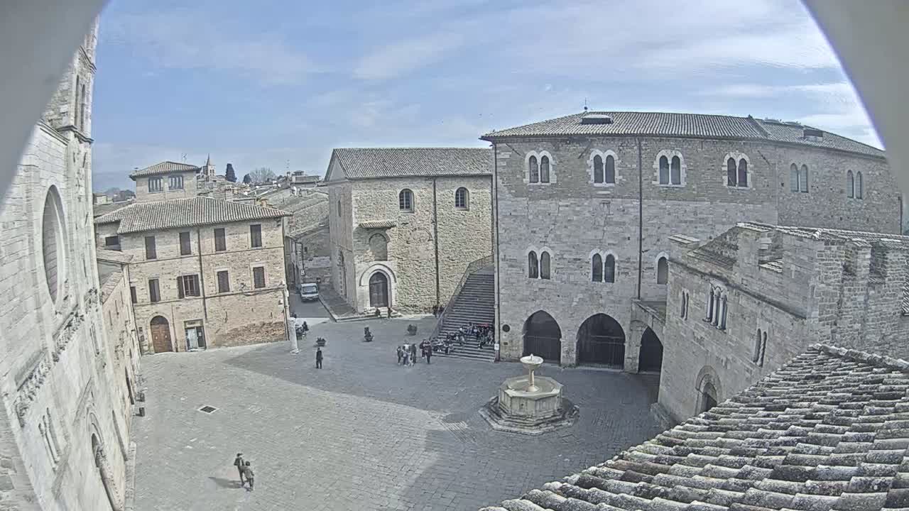 A town square with several stone buildings, a fountain, and a few people, under a clear, sunny sky.
