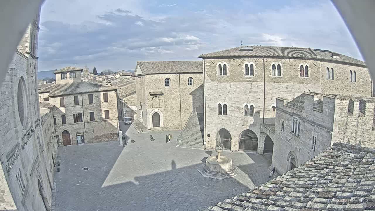 A partly cloudy day reveals an expansive town square surrounded by aged stone buildings, a central fountain, and a few pedestrians.