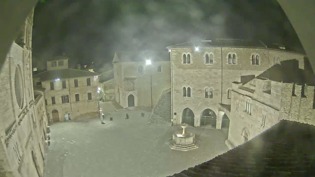 A nighttime view of a town square with several stone buildings, a fountain, and a staircase, under hazy or foggy conditions.