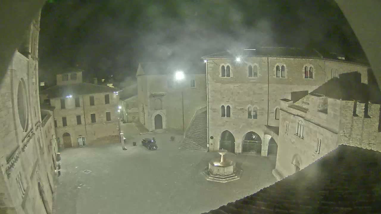 A nighttime view of a town square with several stone buildings, a fountain, and a single car, under a hazy sky.