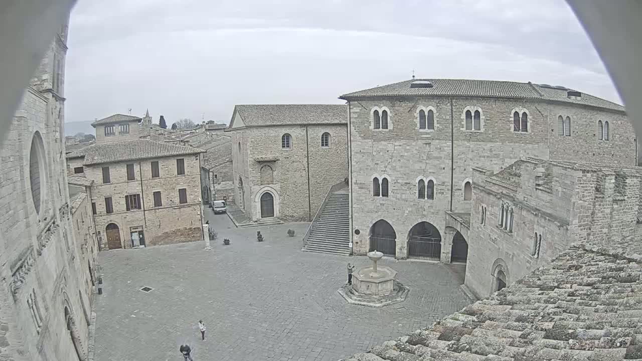 An overcast day reveals a high-angle view of a town square surrounded by aged stone buildings, a central fountain, and a few pedestrians.