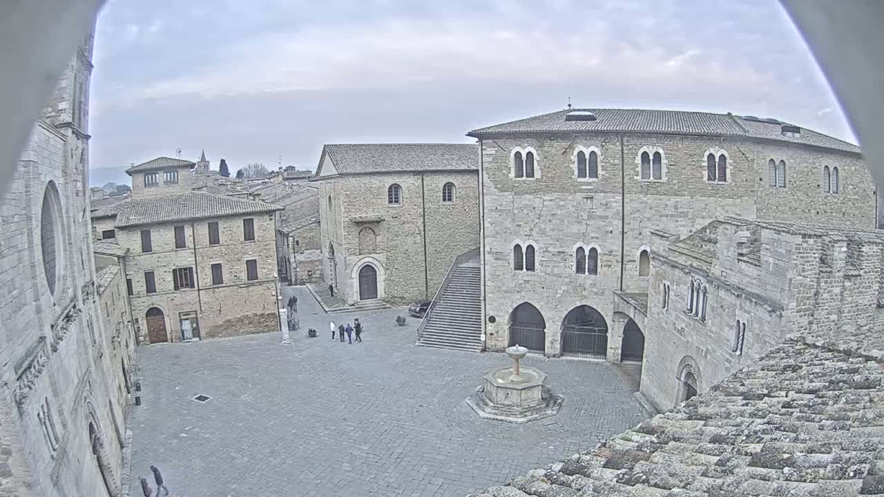 A stone plaza with a fountain is surrounded by medieval-style buildings under an overcast sky.