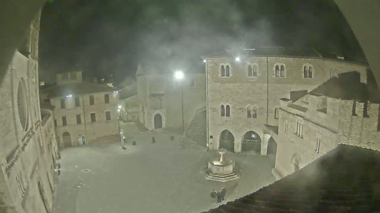 A nighttime view of a town square, shrouded in mist, with several stone buildings surrounding a central fountain.