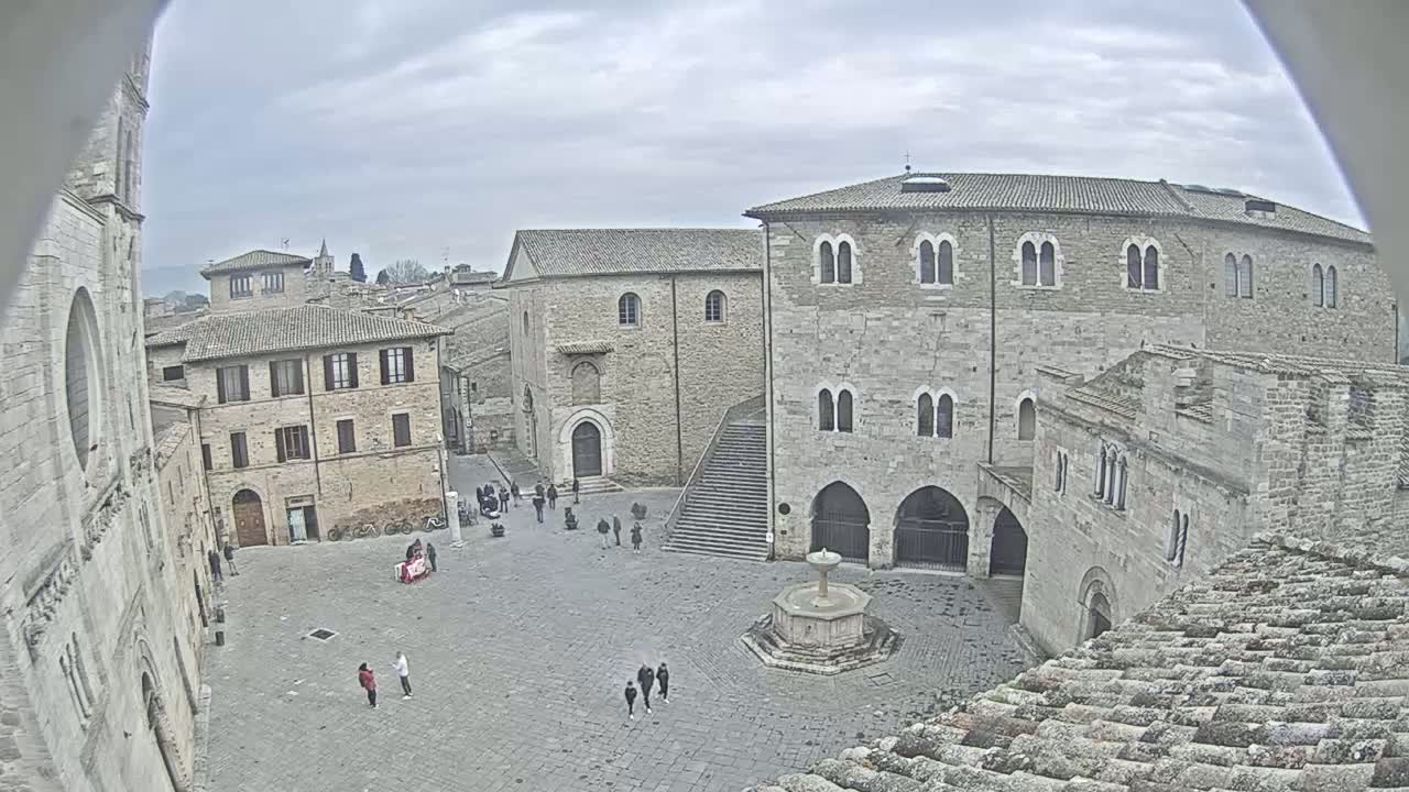 An overcast day reveals a stone town square with several buildings, a fountain, and a few pedestrians.