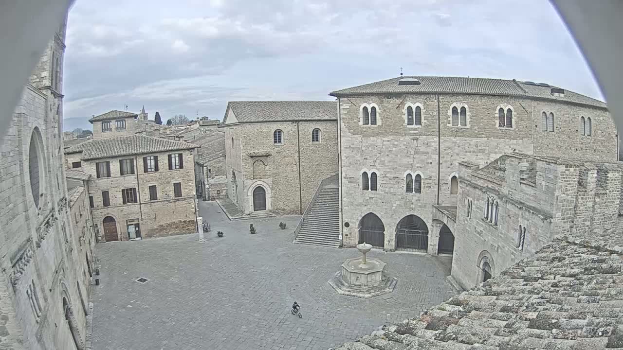 A stone plaza with a fountain and several medieval-style buildings is seen on an overcast day, a single person on a bicycle is visible in the distance.