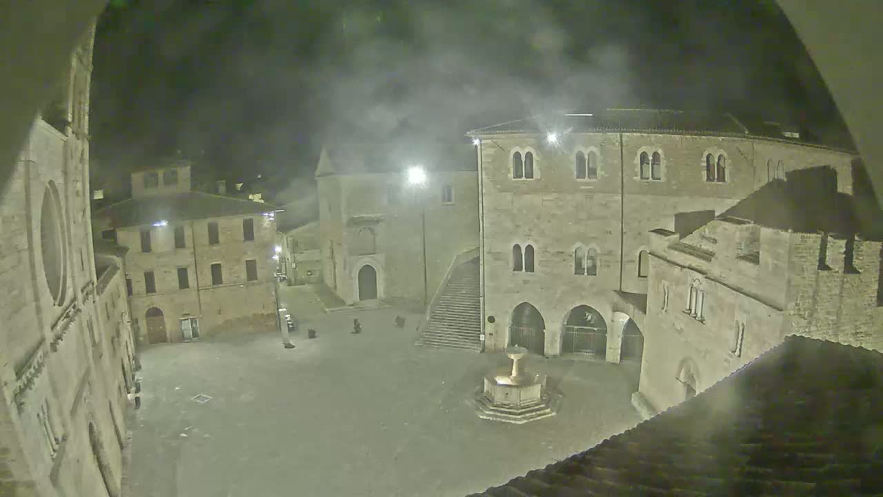 A nighttime, hazy view of a town square surrounded by aged stone buildings, centered on a fountain.