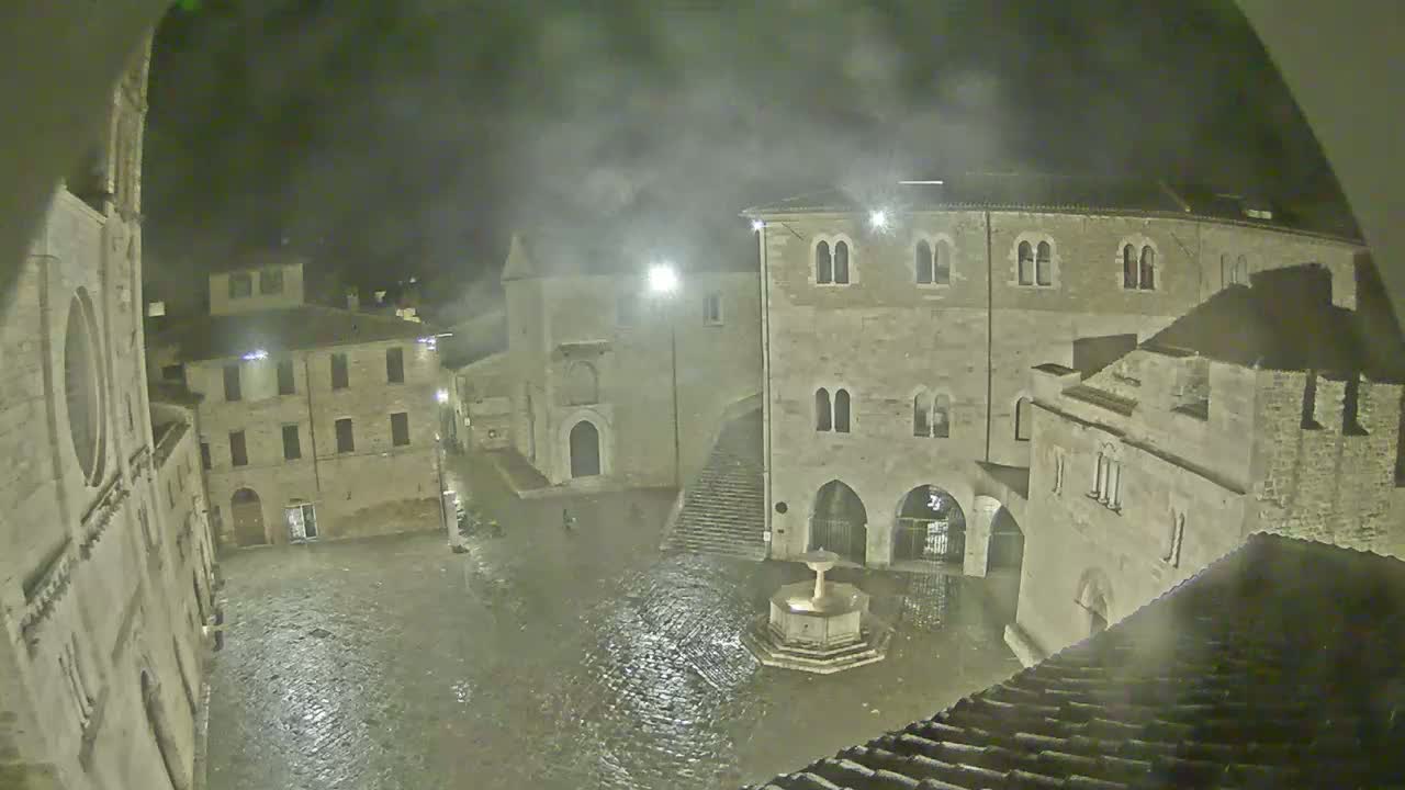 A rain-soaked town square at night, surrounded by old stone buildings and a central fountain.