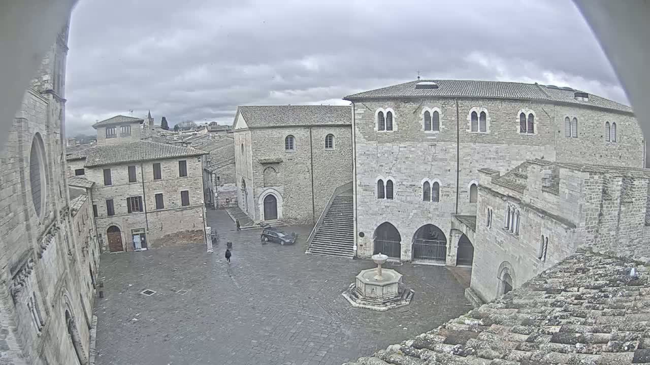 A stone plaza with a fountain is surrounded by ancient-looking buildings under an overcast sky.