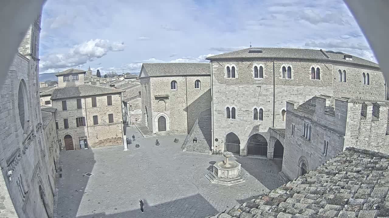 A mostly empty stone plaza in a historic town, viewed from above on a partly cloudy day, features several ancient buildings surrounding a central fountain.