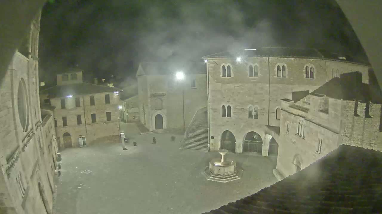 A nighttime view of a town square with stone buildings, a fountain, and hazy atmospheric conditions.