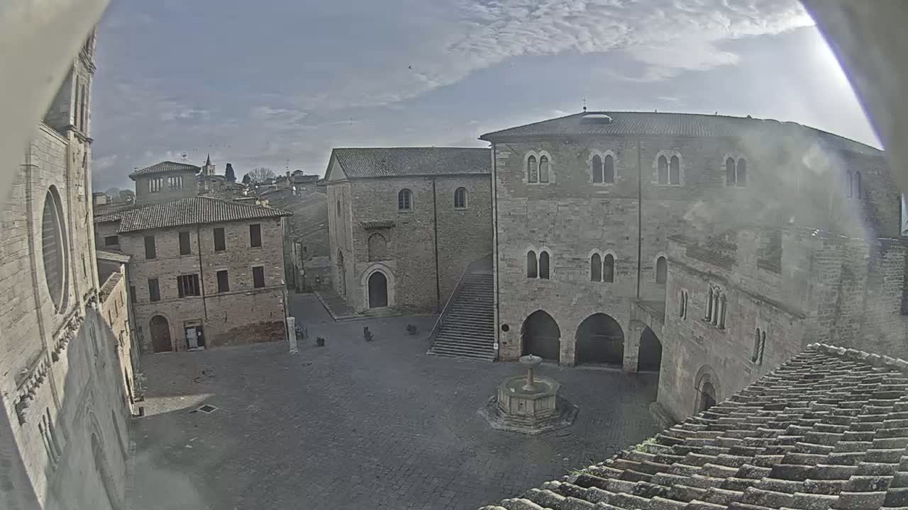 A mostly cloudy day reveals an open square surrounded by aged stone buildings, one with a fountain at its center.