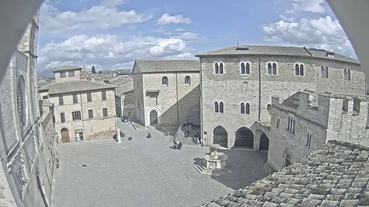 A partly sunny day reveals a town square in a historic European town, surrounded by stone buildings, including a large, imposing structure and a smaller church-like building, with a fountain in the center.