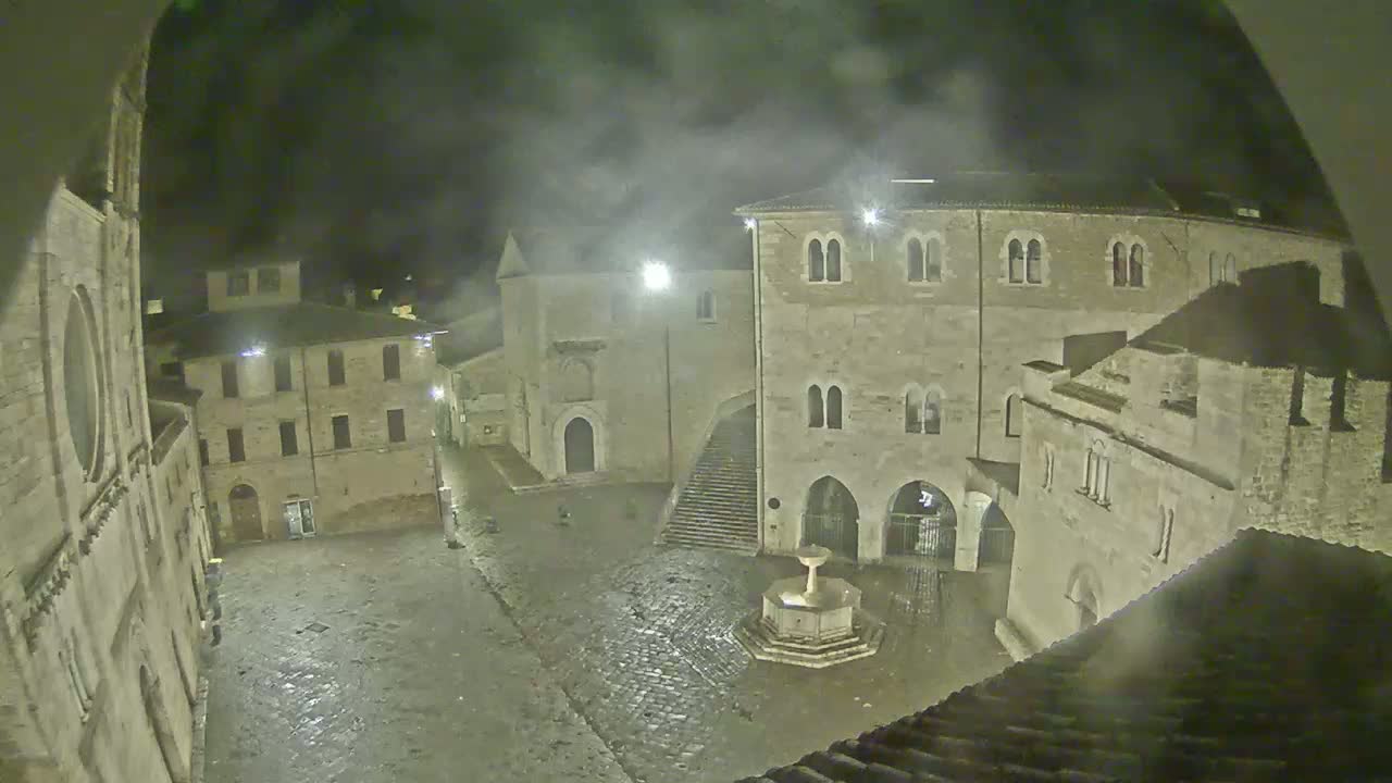 A nighttime view of a cobblestone town square with stone buildings surrounding it, a fountain in the center, and a misty or rainy atmosphere.