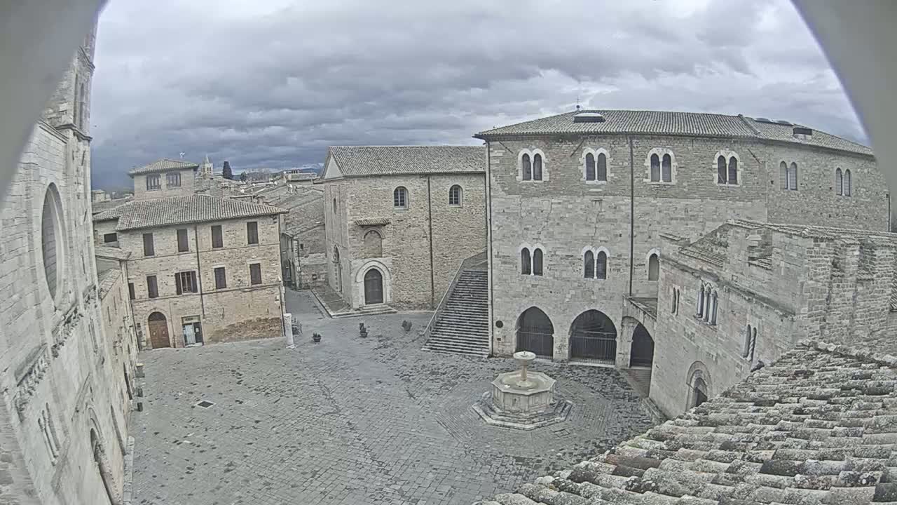 A stone plaza with a fountain, surrounded by medieval-style buildings under a cloudy sky.