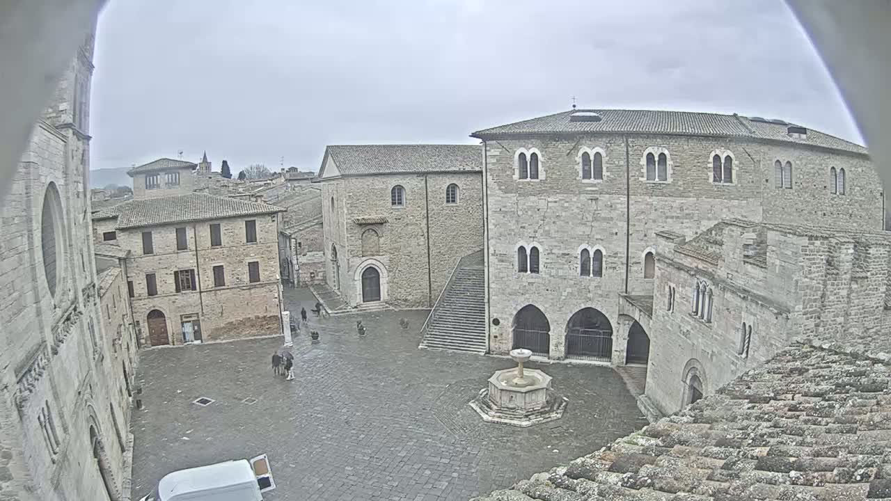 A stone plaza with a fountain is surrounded by medieval-style buildings under an overcast sky.
