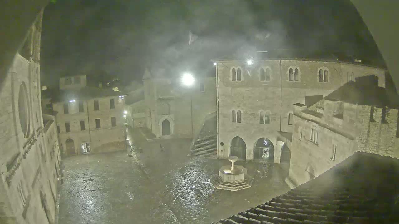 A rain-soaked town square at night, centered around a fountain, is surrounded by ancient stone buildings under a hazy sky.