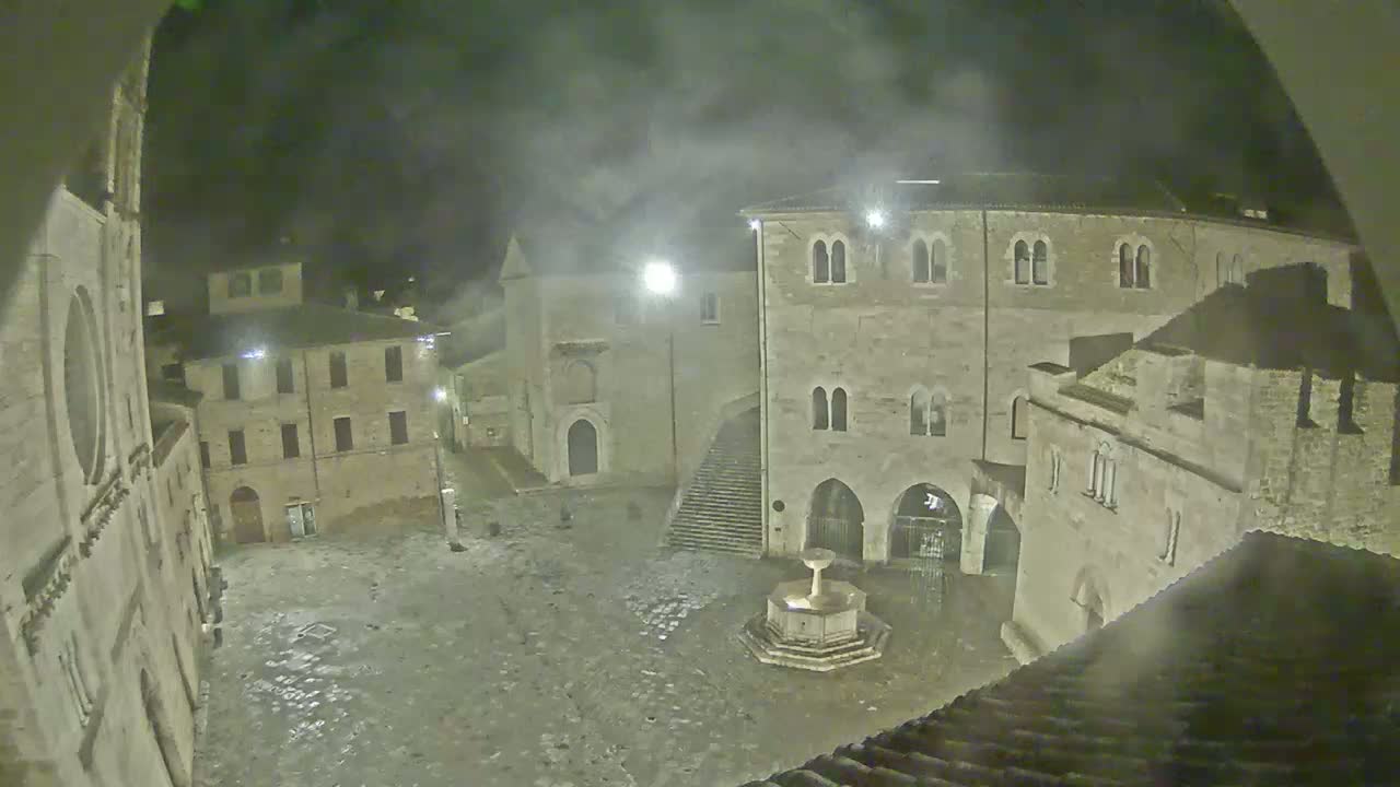 A stone plaza at night, under a hazy sky, is centered around a fountain, surrounded by medieval-style buildings.