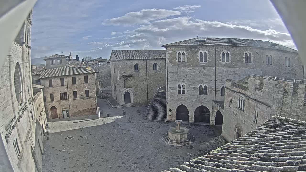A mostly sunny day reveals a stone plaza in a historic town, surrounded by aged buildings and a fountain at its center.