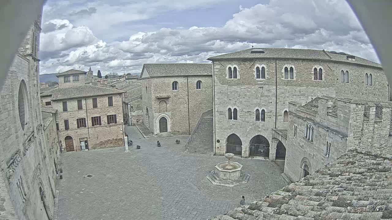 A mostly cloudy sky overlooks a town square with several stone buildings surrounding a fountain, some featuring arched entryways and multiple windows.