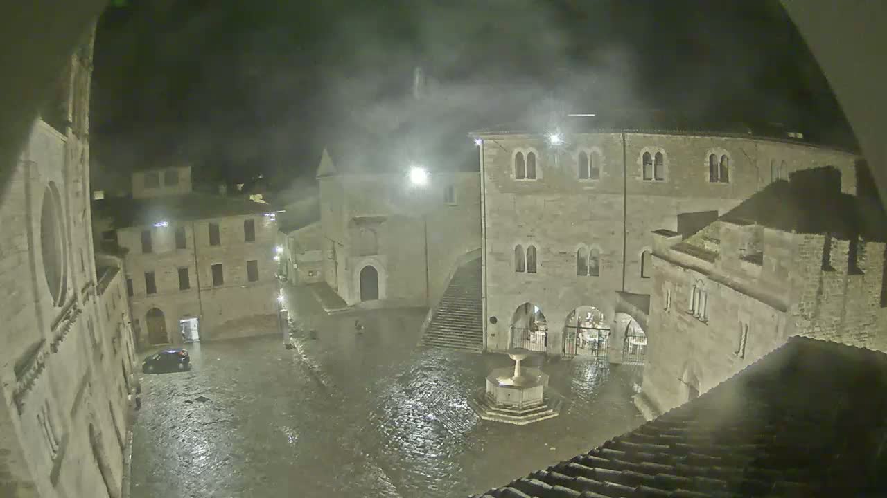 A rain-soaked town square at night, surrounded by ancient stone buildings, features a central fountain.