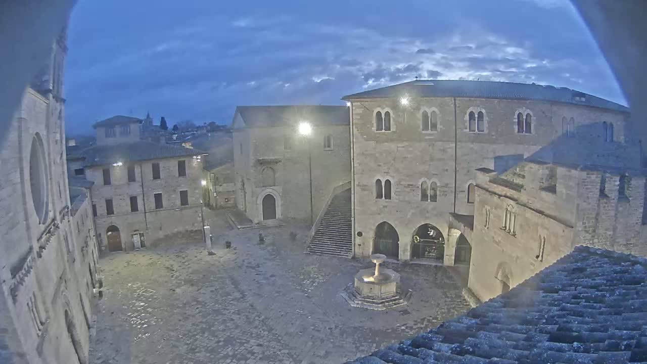 A stone plaza at twilight, centered by a fountain, is surrounded by ancient buildings under a cloudy sky.