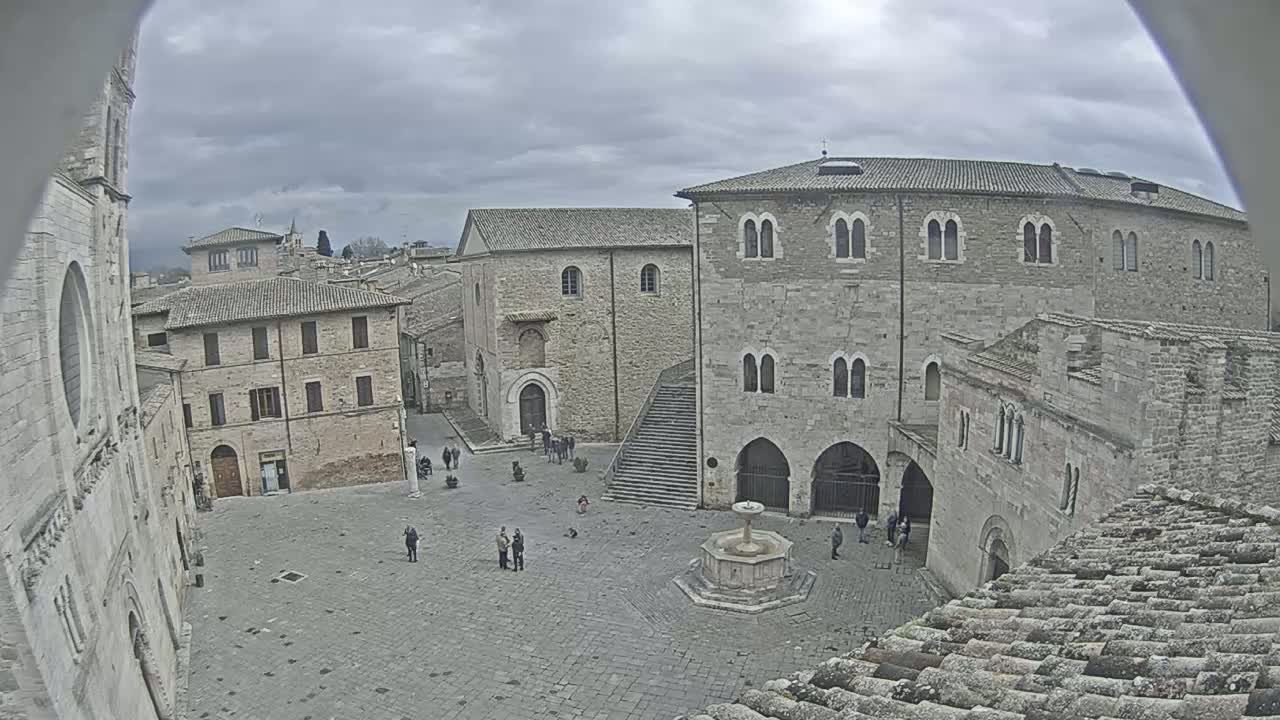 A high-angle view of a town square with several stone buildings surrounding a fountain, under an overcast sky.