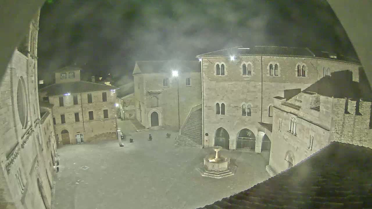 A nighttime view of a town square, with several stone buildings surrounding a central fountain, under hazy or foggy conditions.