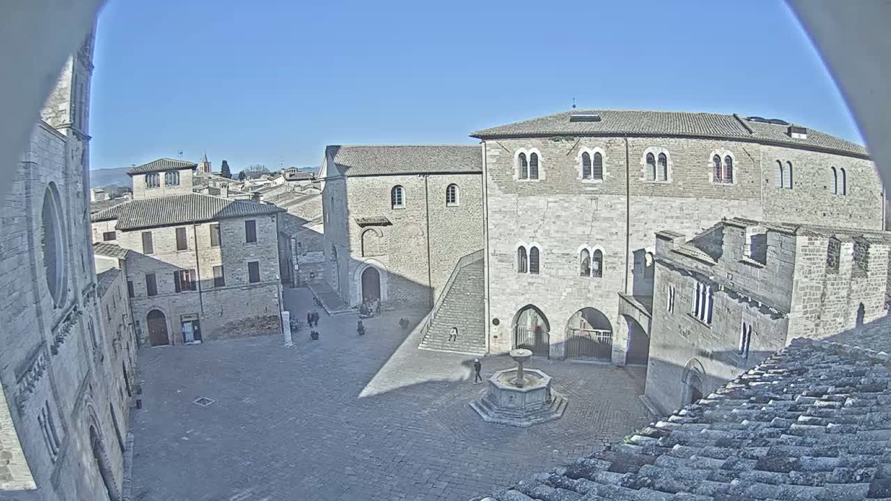 A sunny, high-angle view of a town square, centered around a fountain, surrounded by aged stone buildings.