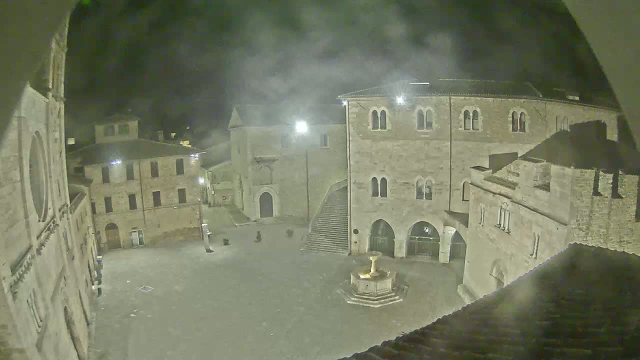 A nighttime, hazy view of a town square with several stone buildings surrounding a fountain.